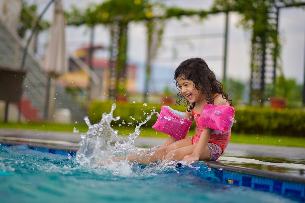 Young girl sitting on the edge of a pool and kicking her feet in the water.